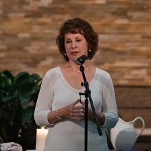 Click to show details - Rev. Meemie Lohmueller | Pastoral Care Minister Meemie Lohmueller wearing white dress standing in front of a microphone in a church sanctuary
