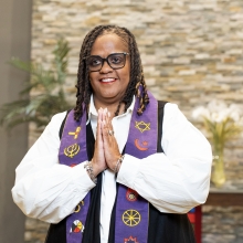 Rev. Tami Johns on stage in church sanctuary wearing white blouse with purple interfaith sash, hands clasped in prayer, smiling.