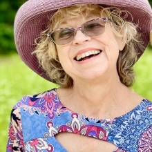 Unity SpiritGroup facilitator Dona McNeill wearing a wide brim light purple had and a flowery dress, smiling while standing in an outside setting