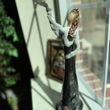 female clay figurine with long blond hair, long black dress with hand gently lifted to the sky