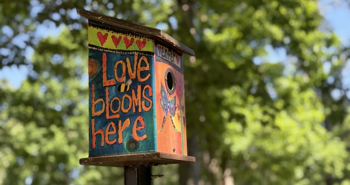 colorful birdhouse with painted words "love blooms here" surrounded by green trees