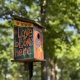 colorful birdhouse with painted words "love blooms here" surrounded by green trees
