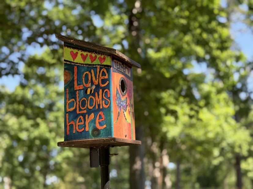 colorful birdhouse with painted words "love blooms here" surrounded by green trees